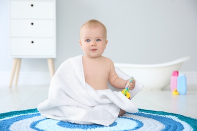 Cute little baby with soft towel and rattle on rug in bathroom