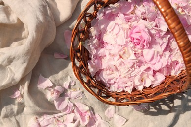 Wicker basket of beautiful tea rose petals on beige fabric, flat lay