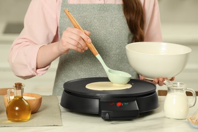 Photo of Woman cooking delicious crepe on electric maker at white marble table in kitchen, closeup
