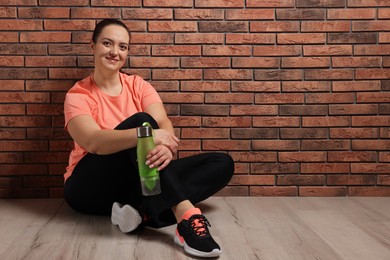Happy overweight woman with bottle of water sitting near brick wall indoors, space for text