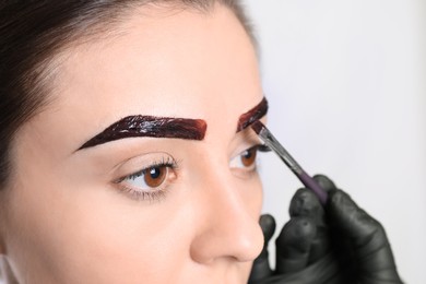 Beautician applying tint during eyebrows correction procedure on light background, closeup