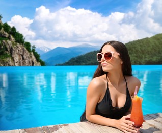 Beautiful young woman with cocktail in outdoor swimming pool at luxury resort and beautiful view of mountains on sunny day