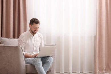 Smiling man holding laptop on armchair near window with beautiful curtains at home. Space for text