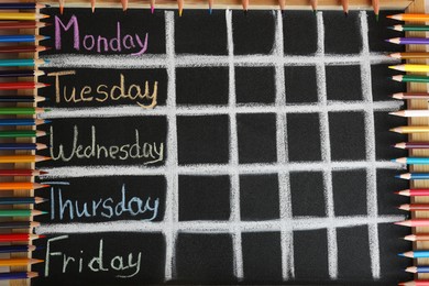 Photo of Colorful pencils and drawn weekly school timetable on black chalkboard, top view