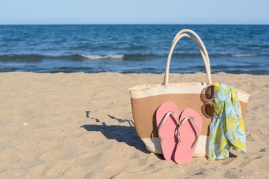 Straw bag with beach wrap, sunglasses and flip flops on sandy seashore, space for text. Summer accessories