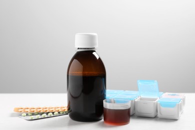 Photo of Bottle of syrup, measuring cup, weekly pill organizer and pills on white table against light grey background. Cold medicine