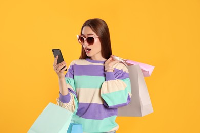 Photo of Surprised young woman with shopping bags looking at smartphone on yellow background. Big sale
