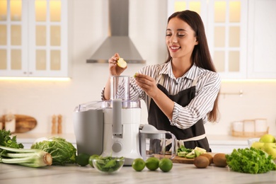 Young woman putting fresh kiwi and apple into juicer at table in kitchen