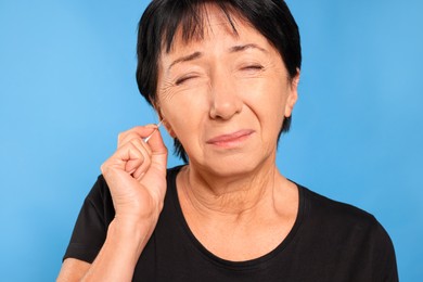 Photo of Senior woman cleaning ear with cotton swab on light blue background, closeup