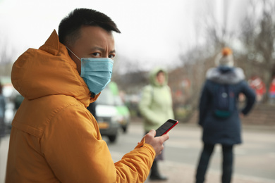 Asian man with smartphone wearing medical mask on city street, space for text. Virus outbreak