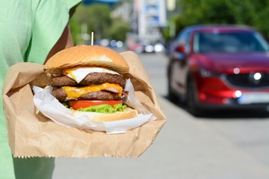 Photo of Woman holding delicious burger in paper wrap on city street, closeup. Space for text
