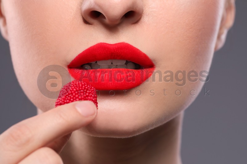 Young woman with beautiful red lips makeup eating candy on grey background, closeup