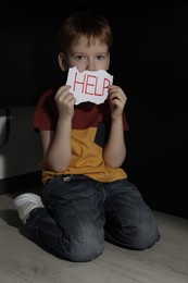 Photo of Scared little boy holding piece of paper with word Help under table on floor indoors. Domestic violence concept