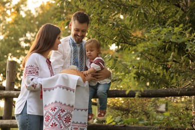 Photo of Happy cute family in embroidered Ukrainian shirts with korovai bread near rustic fence outdoors. Space for text