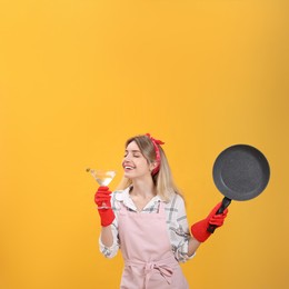 Young housewife with frying pan and glass of martini on yellow background
