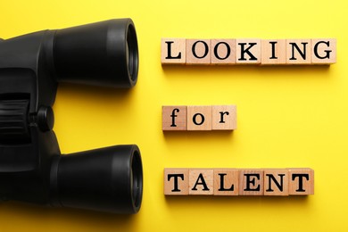 Photo of Staff recruitment concept. Phrase Looking For Talent made of wooden cubes and binoculars on yellow background, flat lay