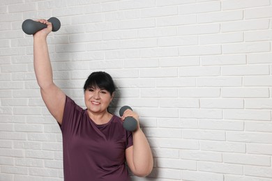 Happy overweight mature woman doing exercise with dumbbells near white brick wall, space for text