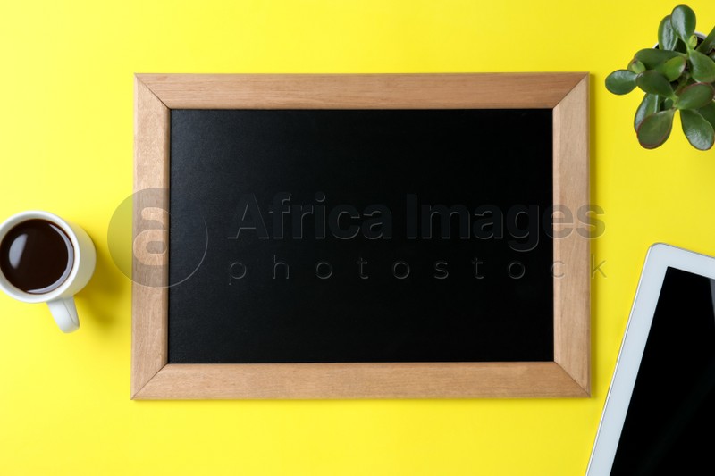 Clean small chalkboard, coffee, plant and tablet on yellow background, flat lay
