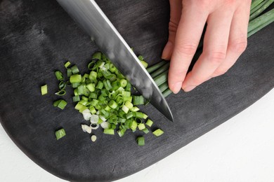 Woman cutting green spring onion on black wooden board at white table, closeup