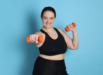 Happy overweight woman doing exercise with dumbbells on light blue background