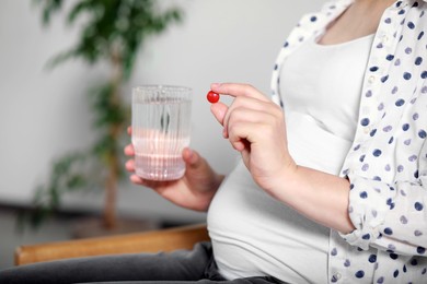 Pregnant woman holding pill and glass of water indoors, closeup