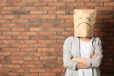 Woman wearing paper bag with drawn sad face near brick wall. Space for text