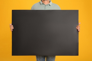 Man holding black blank poster on yellow background, closeup. Mockup for design