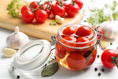 Glass jar of pickled cherry tomatoes and ingredients on white wooden table, closeup