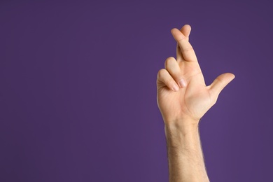 Man with crossed fingers and space for text on purple background, closeup. Superstition concept