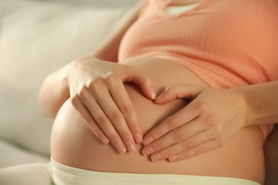 Pregnant woman making heart with her hands near belly indoors, closeup