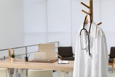 Modern doctor's workplace with wooden table and white coat hanging on rack in office