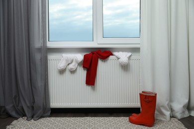 Heating radiator with hat, socks, scarf and rubber boots near window indoors