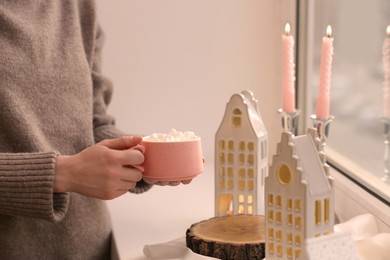 Photo of Woman holding cup of delicious drink near windowsill with house shaped lanterns indoors, closeup. Space for text