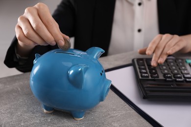 Woman with calculator putting money into piggy bank at grey table, closeup