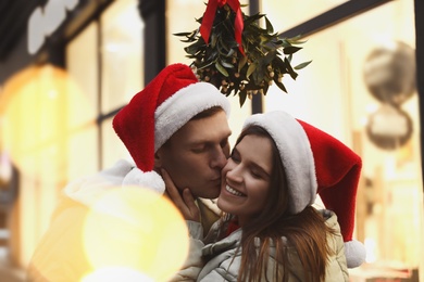 Happy man kissing his girlfriend under mistletoe bunch outdoors, bokeh effect