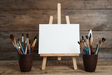 Easel with blank canvas and brushes on wooden table