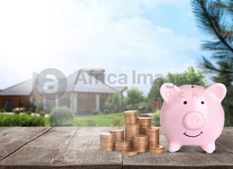 Piggy bank and stacked coins on wooden surface and blurred view of beautiful house, space for text. Mortgage concept