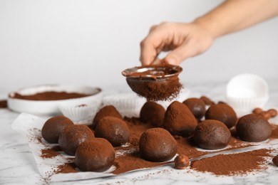 Woman sieving cocoa powder onto delicious chocolate truffles at white marble table, closeup