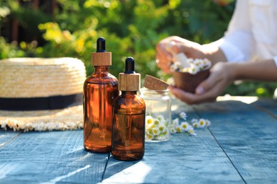 Woman grinding chamomile flowers in mortar outdoors, focus on bottles of essential oil