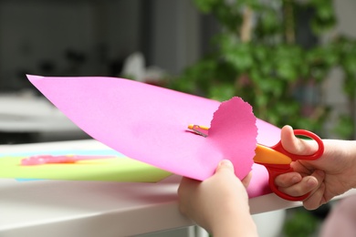 Child cutting out paper heart with craft scissors at table indoors, closeup