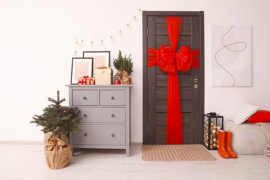 Photo of Beautiful fir tree near chest of drawers and wooden door decorated with red bow in room. Christmas decoration