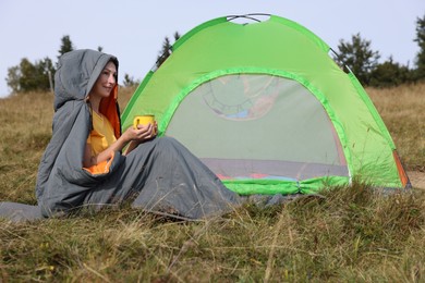 Mature woman with cup of drink in sleeping bag near camping tent on hill