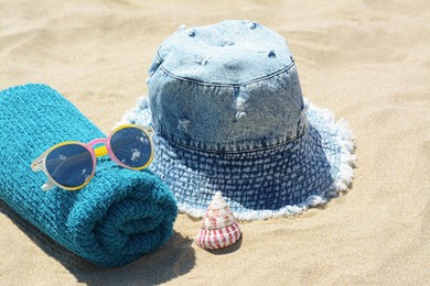 Towel with sunglasses, seashell and denim hat on sand outdoors. Beach accessories
