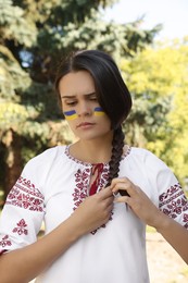 Photo of Sad young woman with drawings of Ukrainian flag on face in park