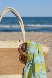 Straw bag with beach wrap and sunglasses on sandy seashore, closeup. Summer accessories