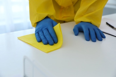 Employee in protective suit and gloves sanitizing table indoors, closeup. Medical disinfection