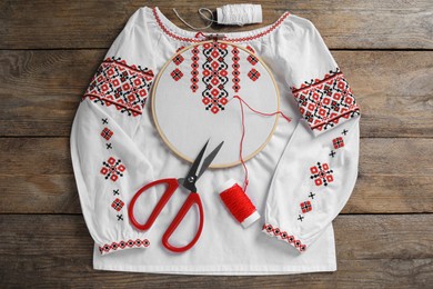 Photo of Shirt with red embroidery design in hoop, needle, scissors and threads on wooden table, flat lay. National Ukrainian clothes