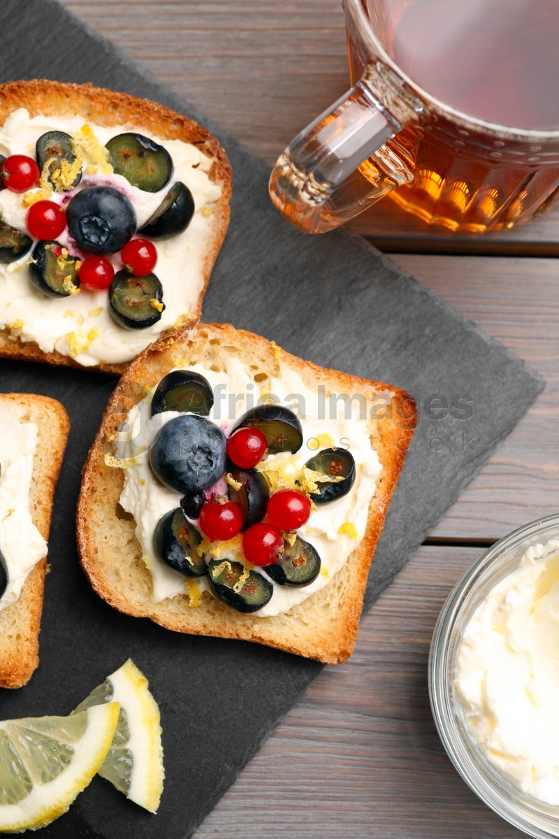 Photo of Tasty sandwiches with cream cheese, blueberries, red currants and lemon zest near cup of tea on wooden table, flat lay