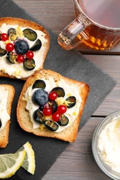 Tasty sandwiches with cream cheese, blueberries, red currants and lemon zest near cup of tea on wooden table, flat lay