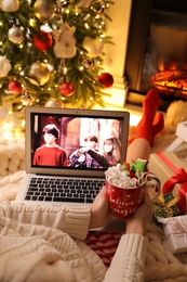 MYKOLAIV, UKRAINE - DECEMBER 23, 2020: Woman with sweet drink watching Harry Potter and Philosopher's Stone movie on laptop near fireplace at home, closeup. Cozy winter holidays atmosphere